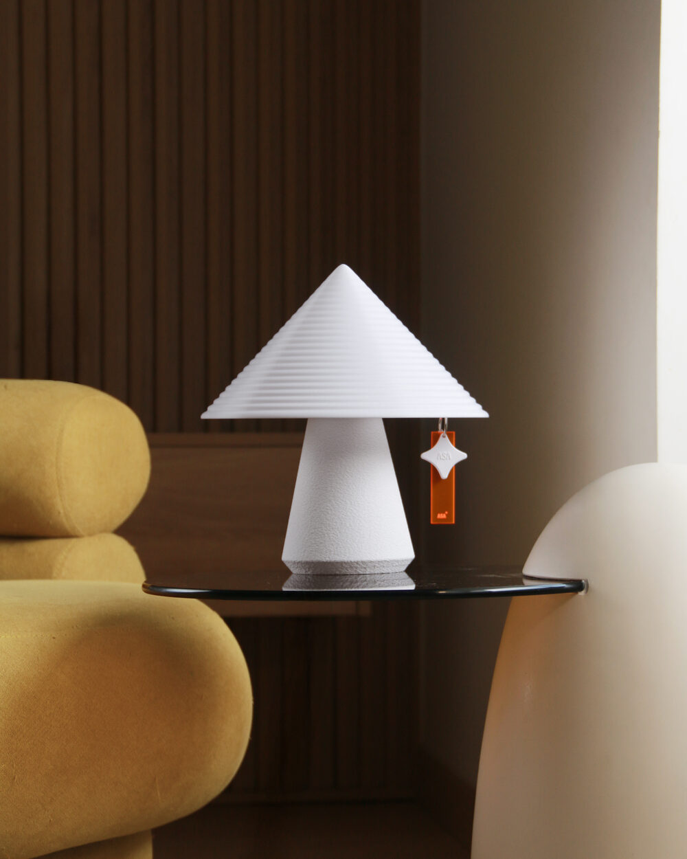 White NOLA lamp with an ASA charm on a glass side table beside a mustard yellow lounge chair