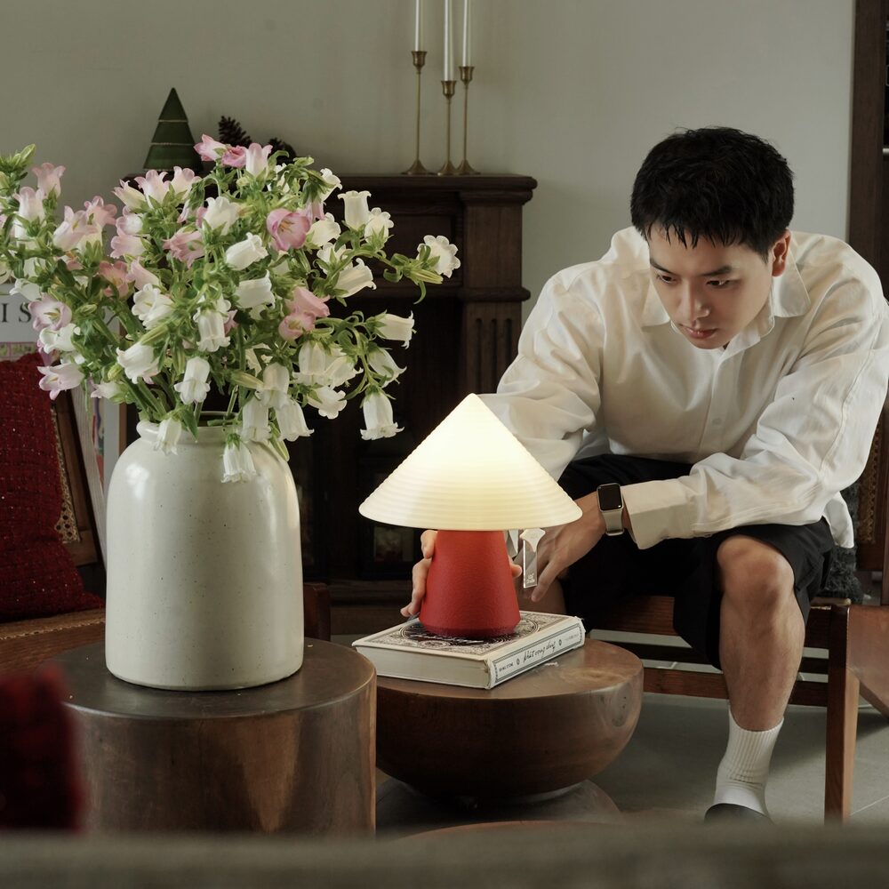 A man adjusting a red NOLA lamp on a stack of books beside fresh flowers in a warm living room
